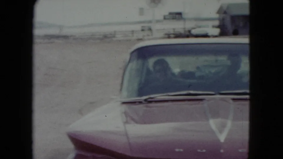 1960 Nevada 8mm frame of a red car near a roadside gas station, with a driver or passenger silhouette visible through the windshield.