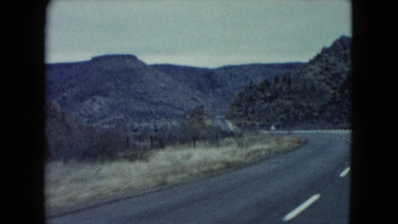 Two-lane highway curves through low mountains under a pale overcast sky.