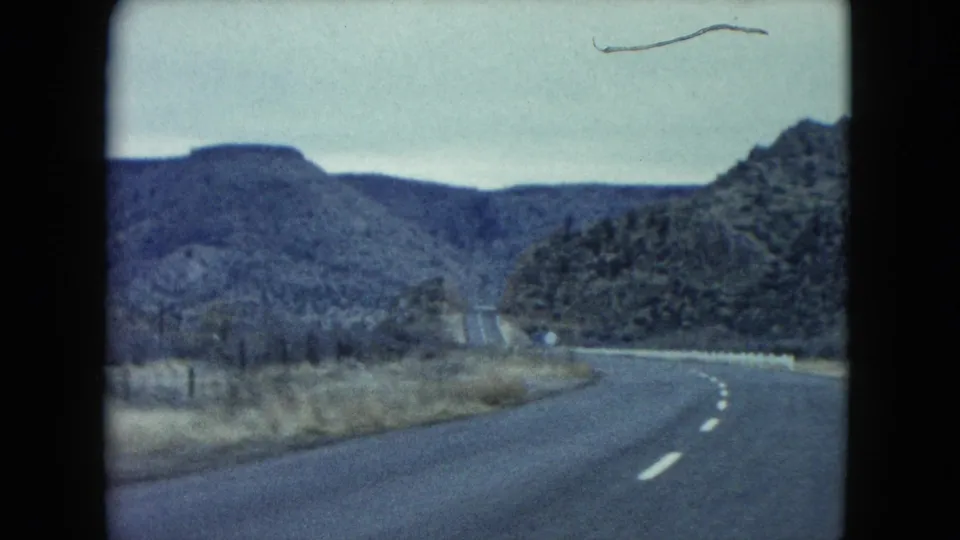 1960 Nevada 8mm view of a winding mountain road, roadside guardrail, scrubland, and a small aircraft-like shape overhead.