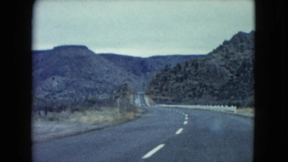 1960 Nevada 8mm frame of a winding highway bordered by scrubland, low mountains, and a light-colored roadside barrier.
