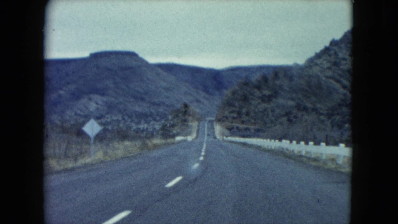 1960 Nevada road-trip frame with a two-lane highway, white guardrail, roadside sign, and rocky hills in the distance.