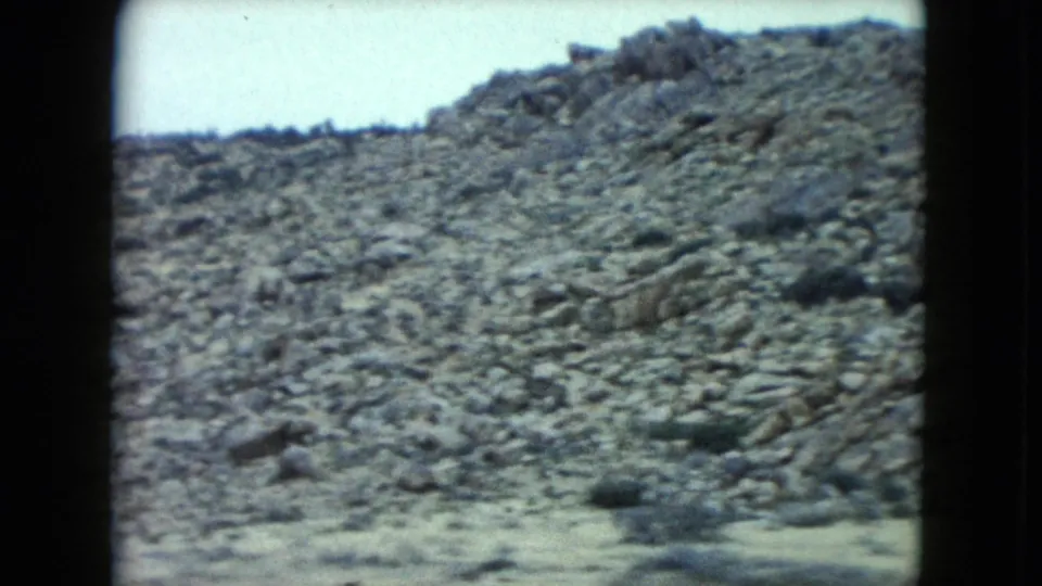 1960 Nevada home-movie frame showing rocky desert ground and low brush passing beside the road.