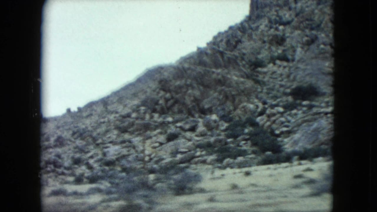 1960 Nevada desert terrain appears through a home-movie frame, with rocky slopes and scattered scrub under pale sky.