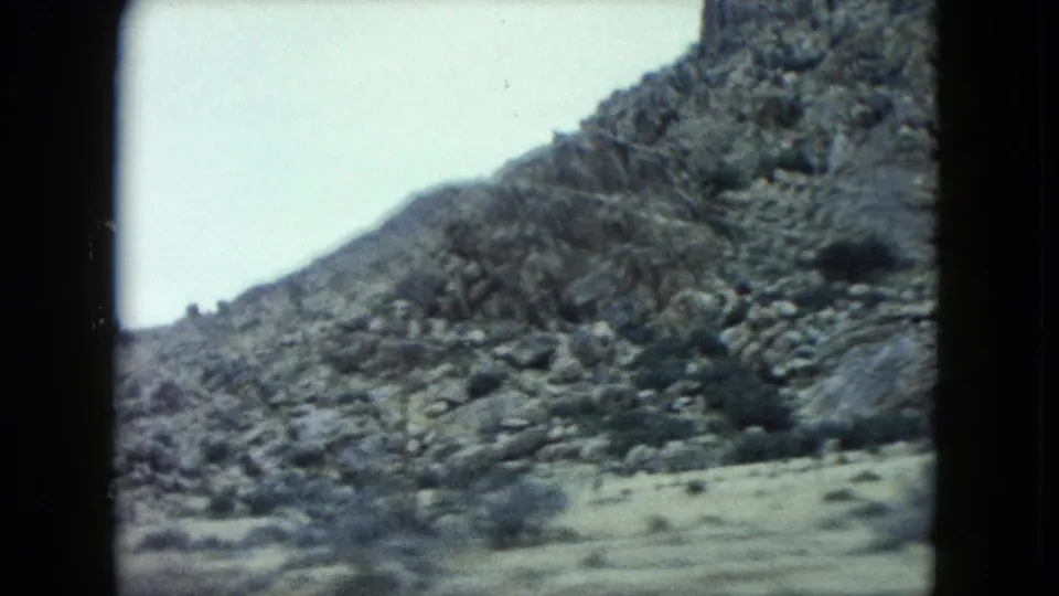 1960 Nevada desert terrain appears through a home-movie frame, with rocky slopes and scattered scrub under pale sky.