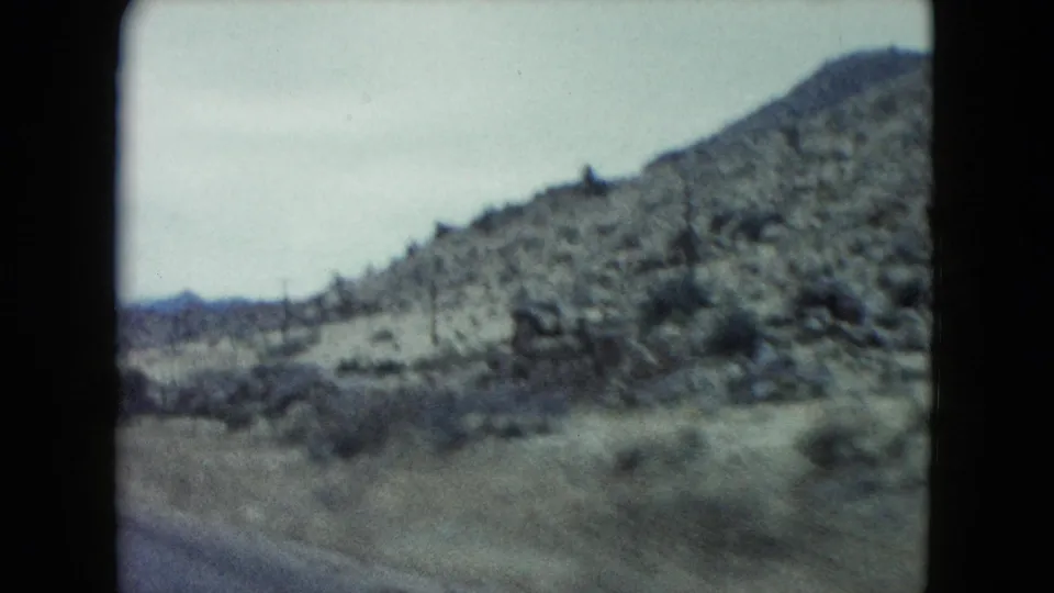 1960 Nevada 8mm travel view with a low desert hill, dry brush, and motion-softened roadside foreground.