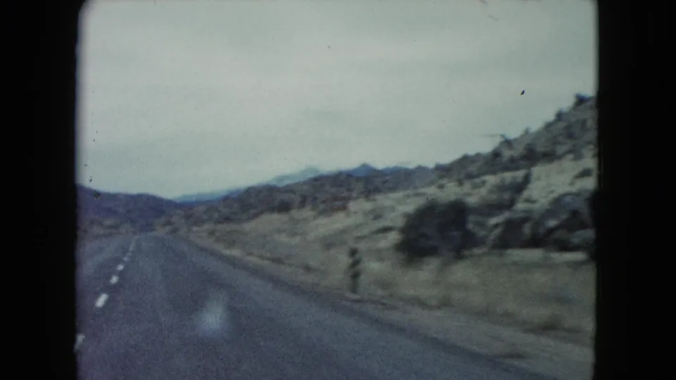 1960 Nevada 8mm frame showing a two-lane road receding through open desert terrain and rocky hills.