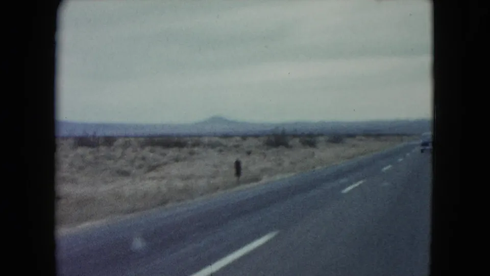 1960 Nevada road-trip frame showing a two-lane highway crossing sparse desert terrain toward distant hills.