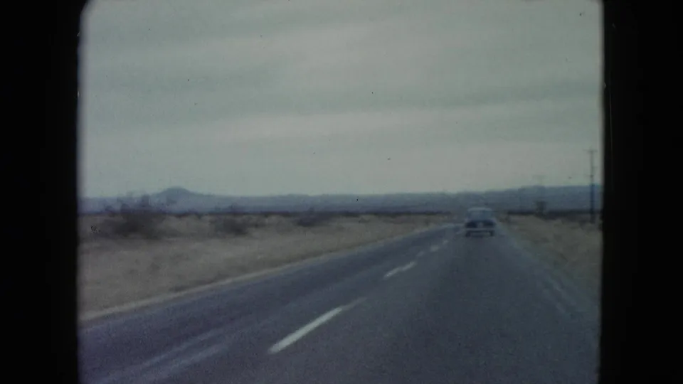 A straight Nevada desert highway in 1960, with a distant car ahead and utility poles along the right side.