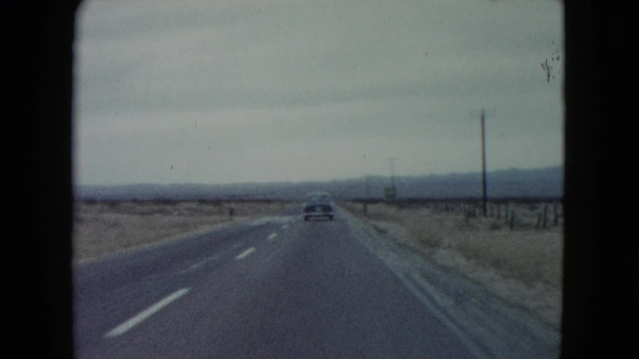 1960 Nevada road-trip view with a single car ahead, roadside poles, wire fencing, and distant desert hills.