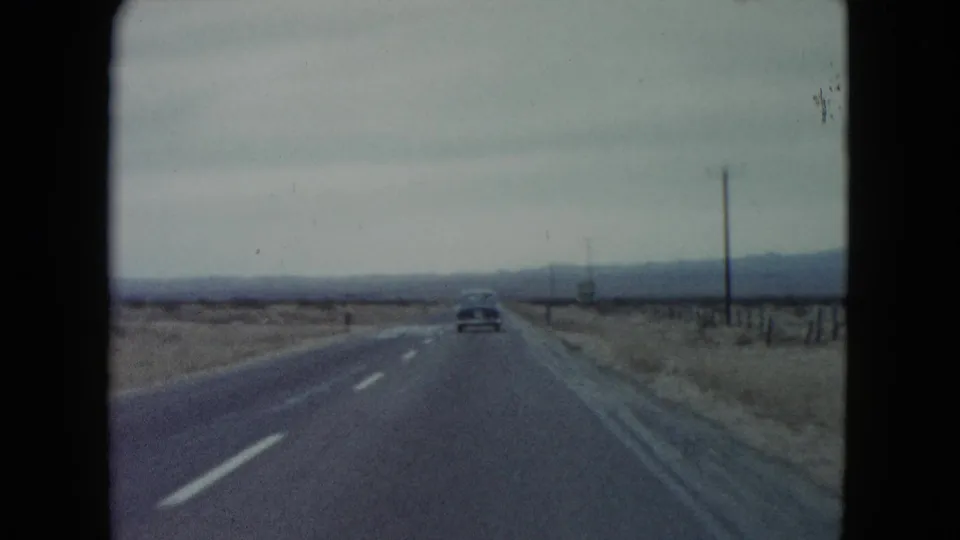 1960 Nevada road-trip view with a single car ahead, roadside poles, wire fencing, and distant desert hills.