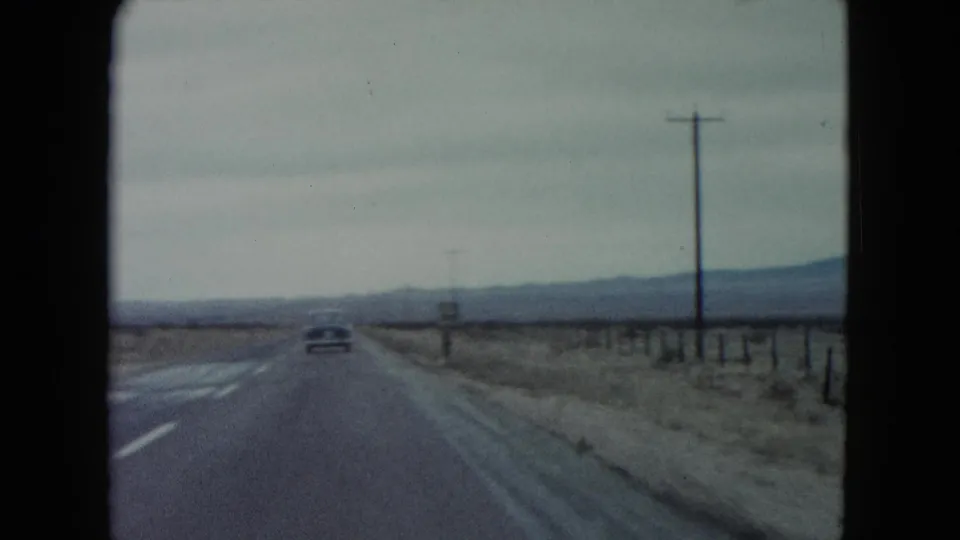 1960 Nevada desert highway frame showing a car ahead near a small roadside sign, poles, fencing, and low mountains.