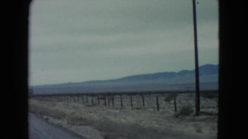 1960 Nevada home-movie frame looking across roadside desert fencing toward low mountains beside the highway.