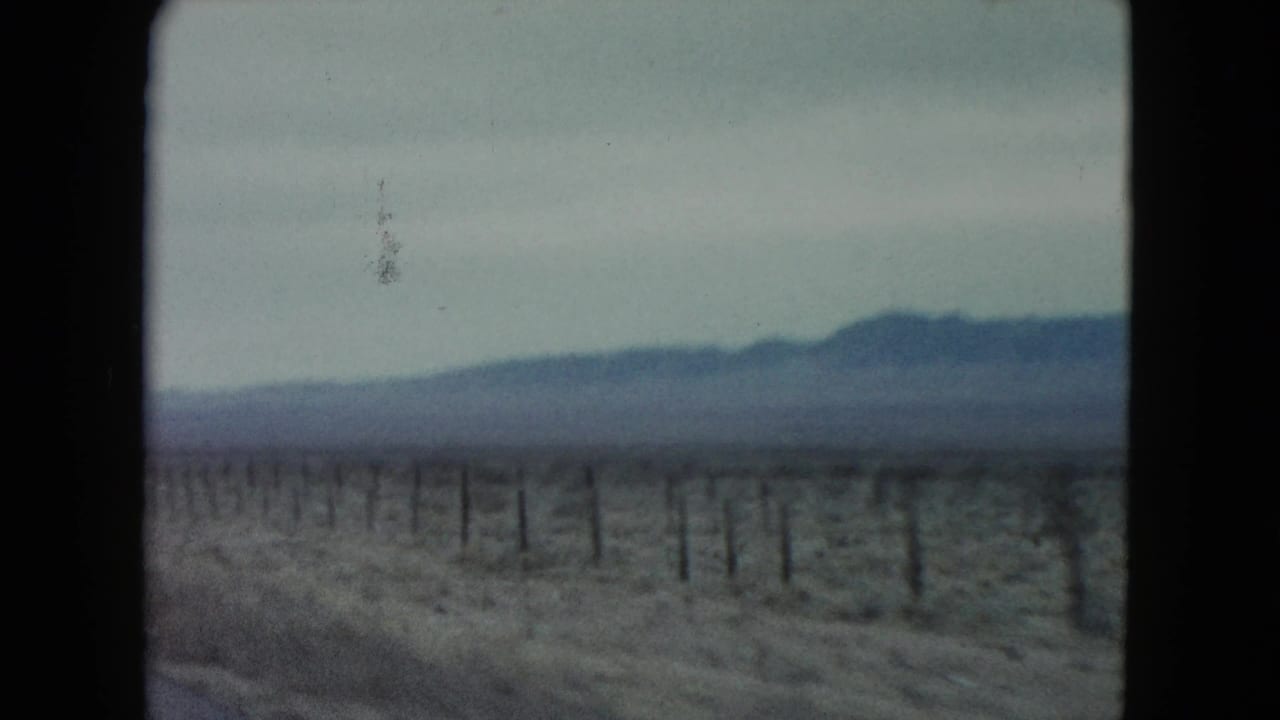 1960 Nevada 8mm frame showing scrubby desert beside a road, a line of fence posts, and distant mountains softened by motion and film grain.
