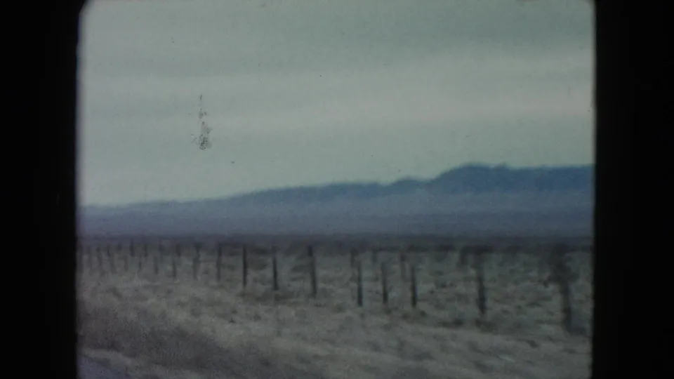 1960 Nevada 8mm frame showing scrubby desert beside a road, a line of fence posts, and distant mountains softened by motion and film grain.