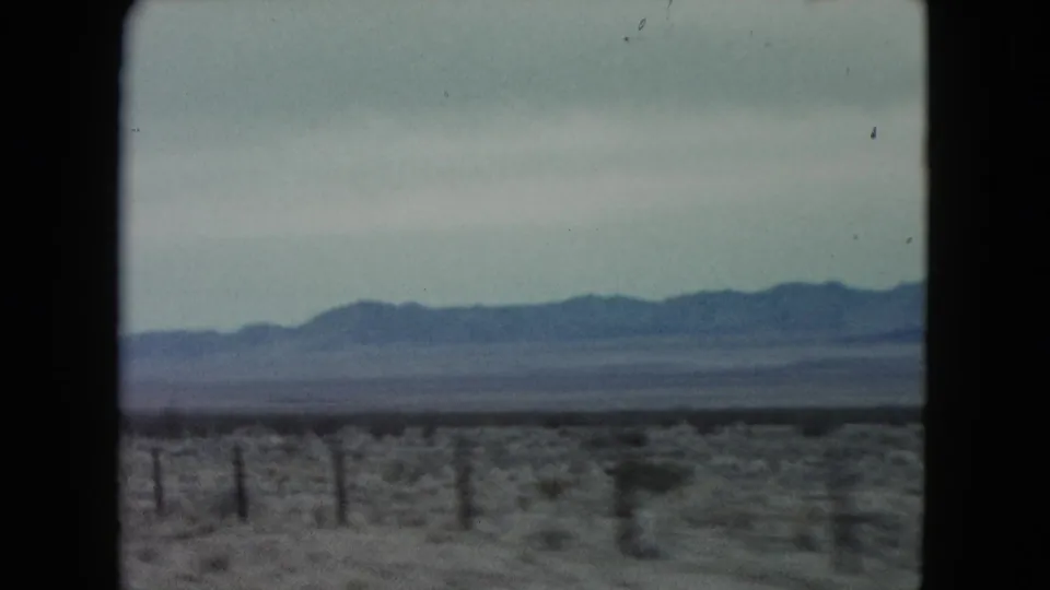 1960 Nevada road-trip view from a moving vehicle, with low desert vegetation, roadside fencing, and a mountain range beneath a muted sky.
