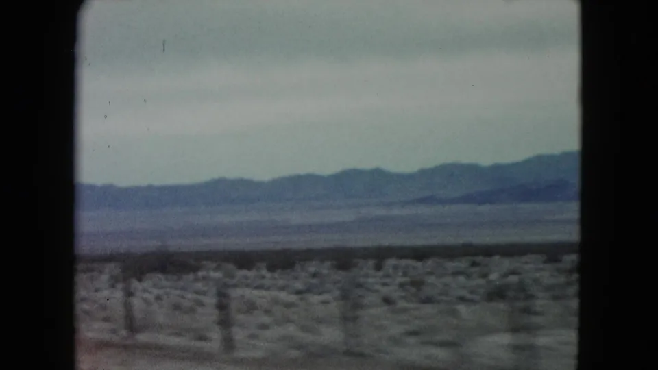Soft 8mm frame from a moving car in Nevada, showing a blurred roadside foreground, scattered fence posts, and a blue mountain ridge across the horizon.