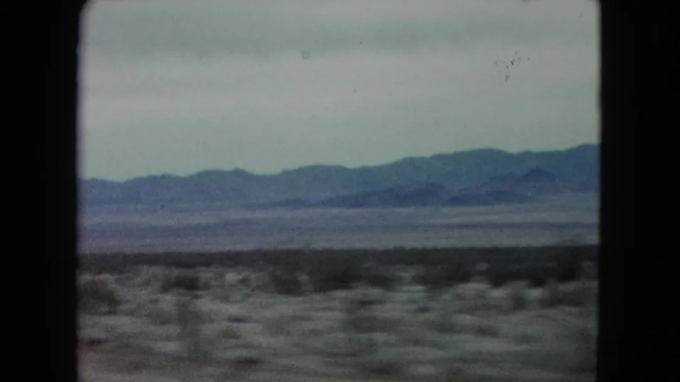 1960 Nevada 8mm travel frame with scrub-covered desert, a dark horizontal band of roadside vegetation, and distant mountains beneath an overcast-looking sky.