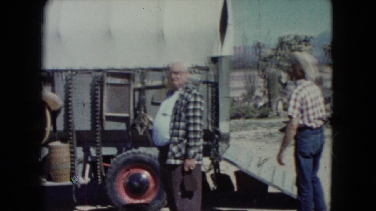 Two men stand near a wagon wheel and ramp in a sunny outdoor western setting.