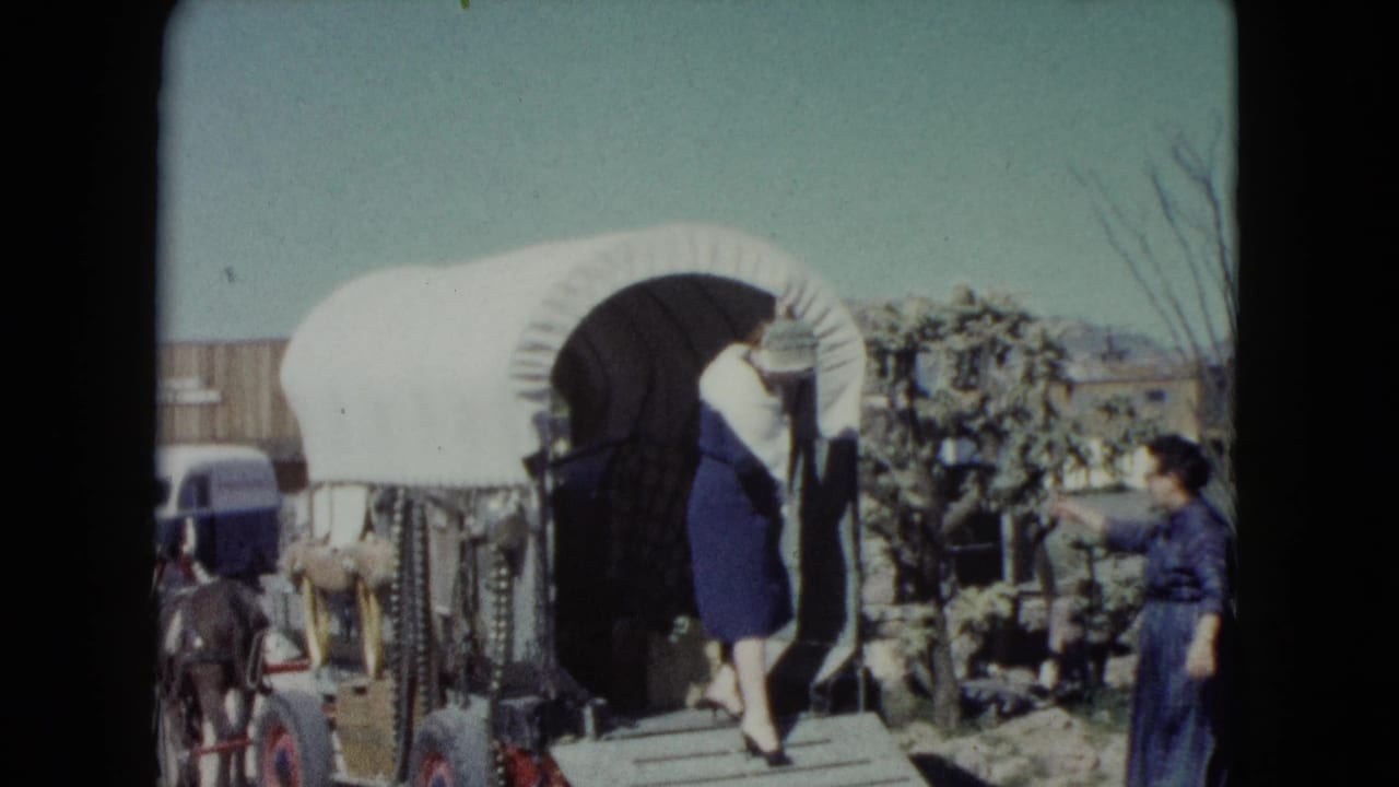 A woman in a blue dress and white shawl steps from a covered wagon as another woman waits nearby in the Nevada desert setting.