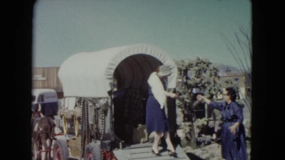 Two women in blue dresses interact beside a covered wagon, with one reaching to assist the other on the wooden ramp.