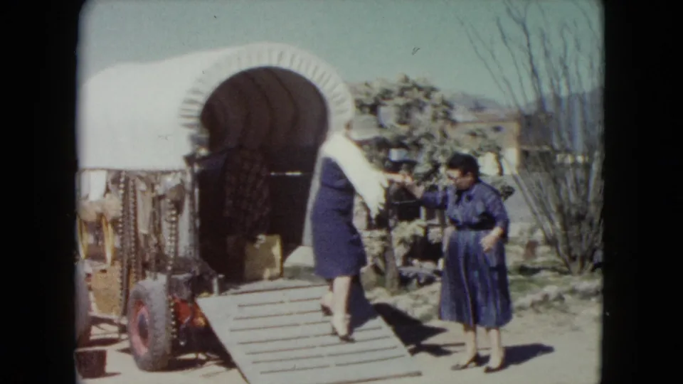 A woman in a white shawl descends the covered wagon ramp as another woman in blue reaches out in the 1960 Nevada home-movie scene.