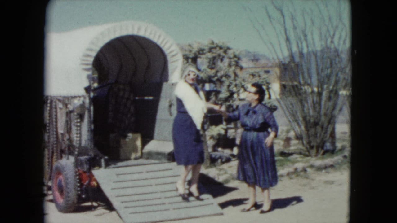 Two women stand by a covered wagon ramp in bright sunlight.