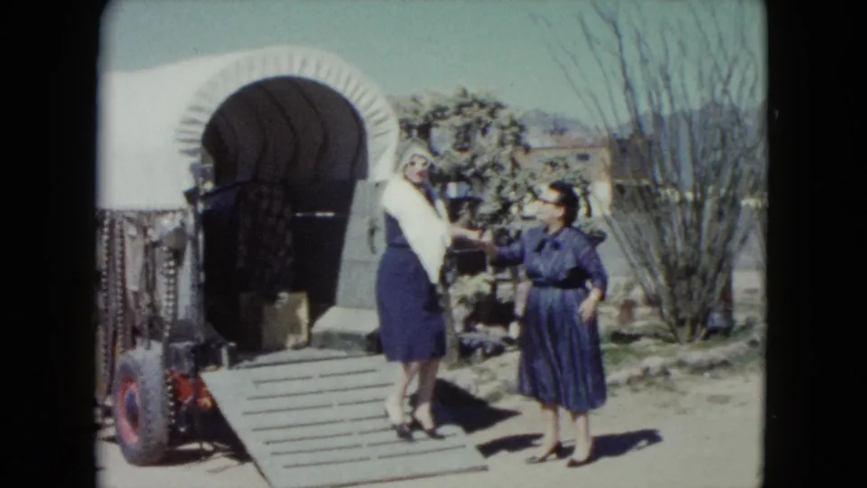 Two adult women in 1960s-style dresses stand beside a white-covered wagon at a Nevada desert roadside setting.