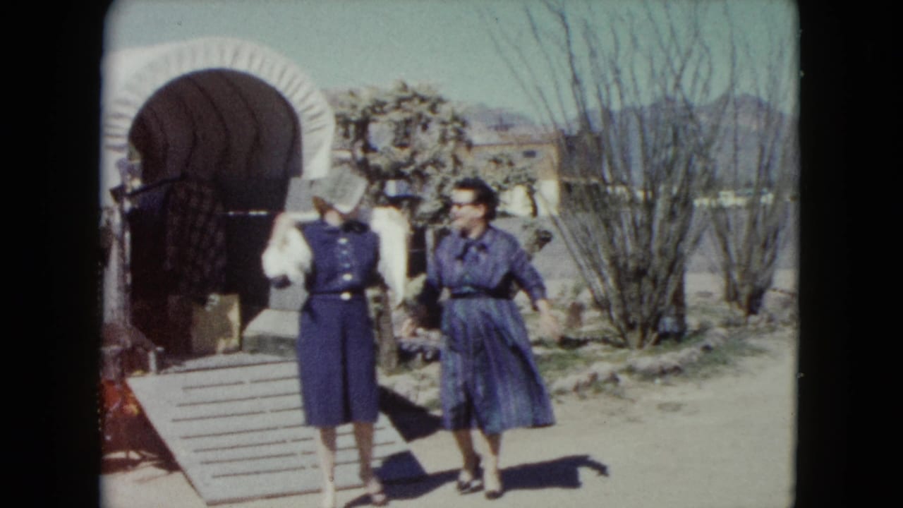 Two adult women in hats and blue dresses move away from a covered wagon in a sunny Nevada roadside scene.