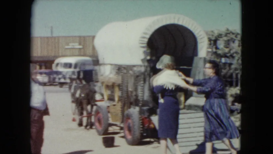 At the covered wagon, one woman with a white shawl stands near the wheel while another woman in a blue dress reaches toward her.