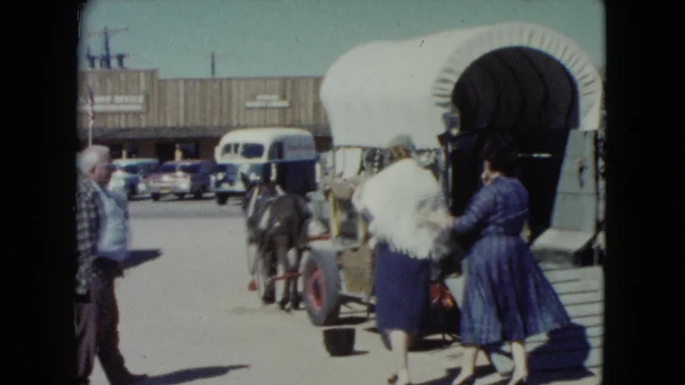 Two adult women remain beside the covered wagon while an onlooker, vintage cars, storefront, and horse-drawn cart appear in the background.