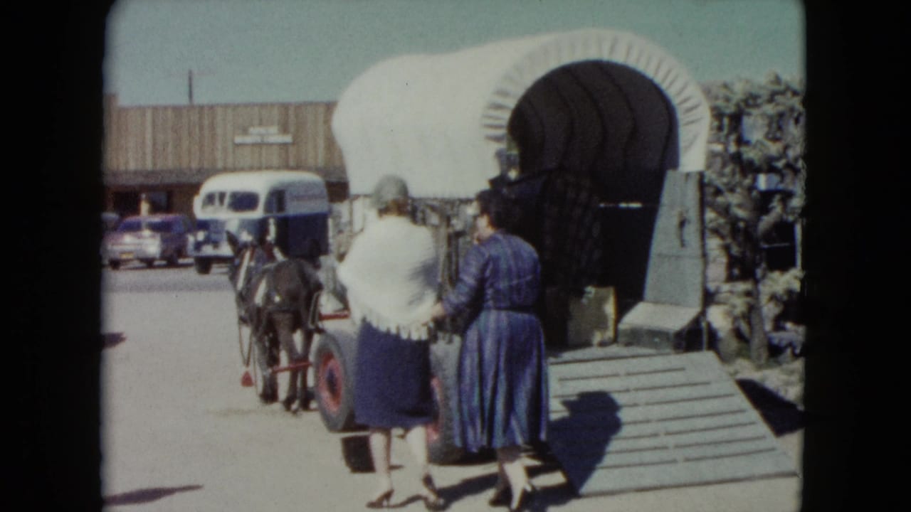 In 1960 Nevada home-movie footage, two women walk toward a mule-drawn covered wagon outside a themed roadside attraction.