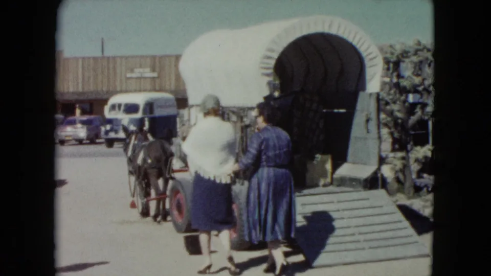 In 1960 Nevada home-movie footage, two women walk toward a mule-drawn covered wagon outside a themed roadside attraction.