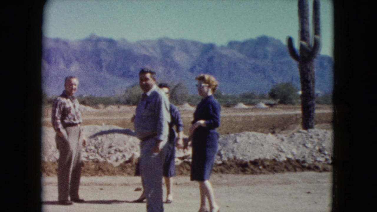 Adults stand talking in a desert area with mountains and a tall cactus behind them.