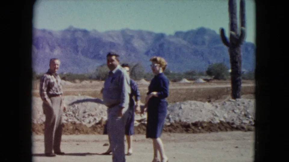 Adult tourists converse in a Nevada desert setting, with rugged mountains and a tall cactus visible behind the group.