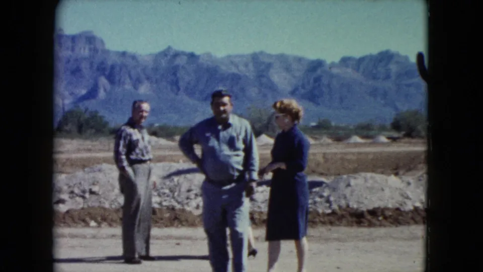 Three adult visitors stand and gesture in a 1960 Nevada desert tourist setting, backed by rocky mountains and open sky.