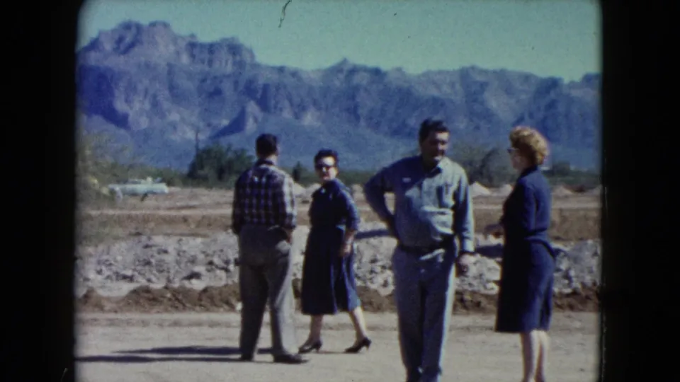 A group of adult tourists stands in the Nevada desert near the attraction, with mountains, cactus, and a small aircraft-like object in the distance.