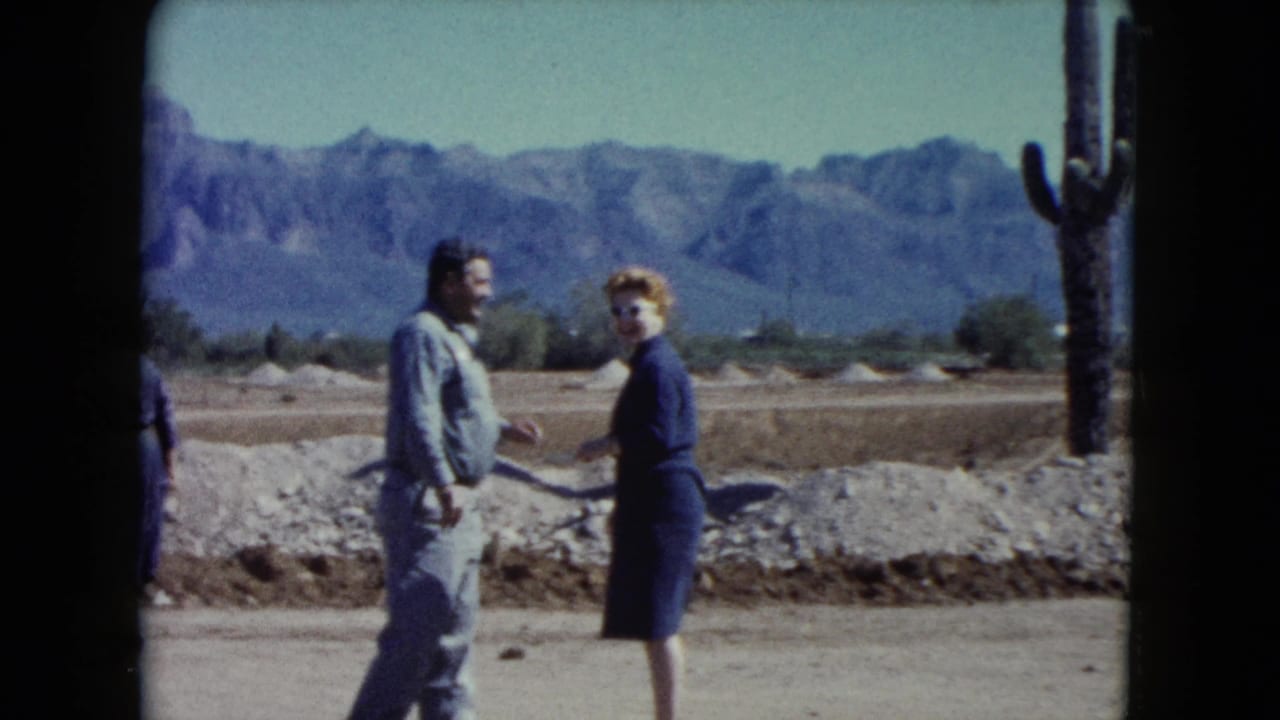 1960 Nevada desert frame with two adults conversing near a tall cactus, rocky ground, open sky, and distant mountains.