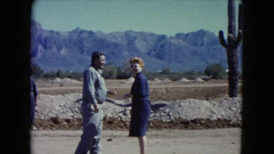 1960 Nevada desert frame with two adults conversing near a tall cactus, rocky ground, open sky, and distant mountains.