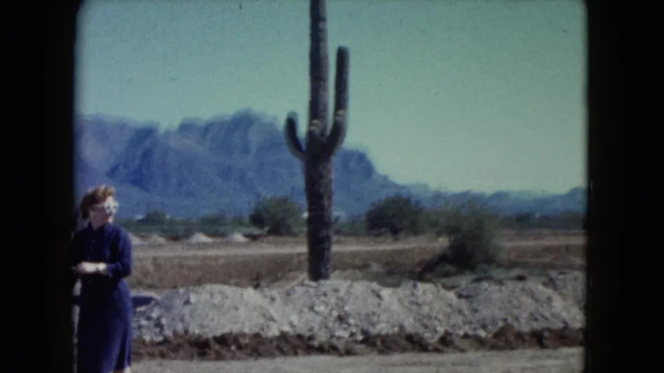 1960 Nevada 8mm frame showing a woman near rocky desert ground, a tall cactus, distant mountains, and pale blue sky.