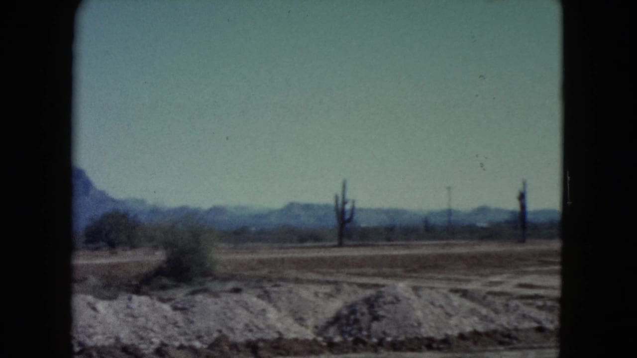 1960 8mm Nevada landscape showing dry desert terrain, scattered brush, cactus forms, and a hazy mountain line.