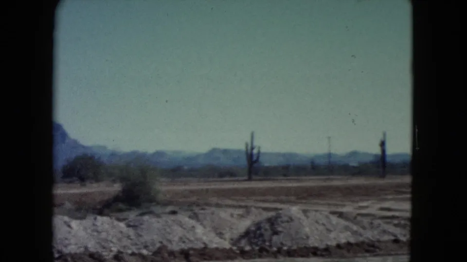 1960 8mm Nevada landscape showing dry desert terrain, scattered brush, cactus forms, and a hazy mountain line.