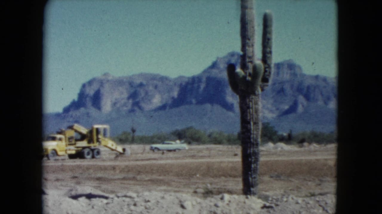 1960 Nevada desert view with a yellow earthmoving vehicle, a period car, cactus, and mountain backdrop.