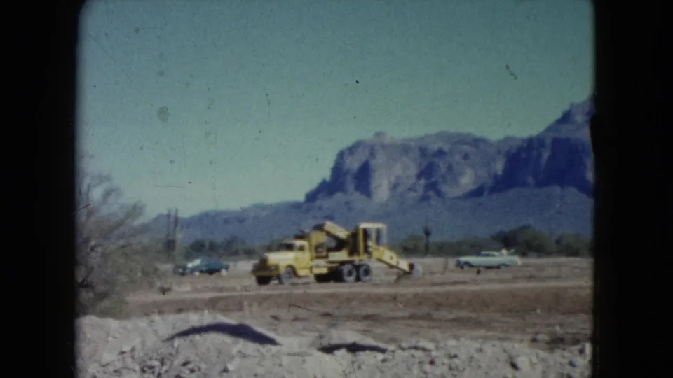 1960 Nevada 8mm frame of roadside construction equipment and cars across open desert terrain below mountains.