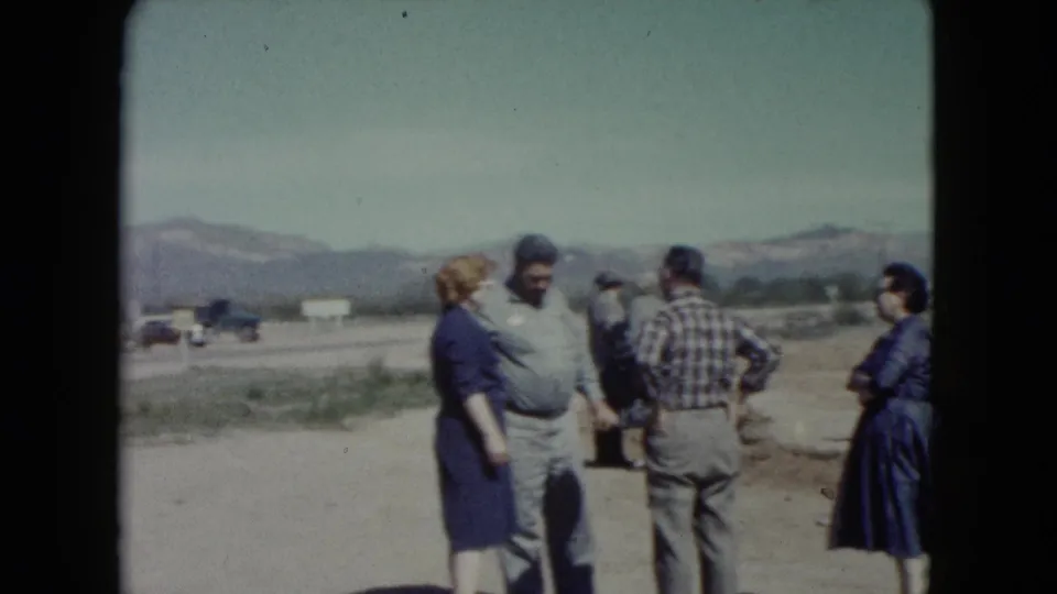 1960 Nevada home-movie frame showing adults standing together near a desert roadside with mountains beyond.
