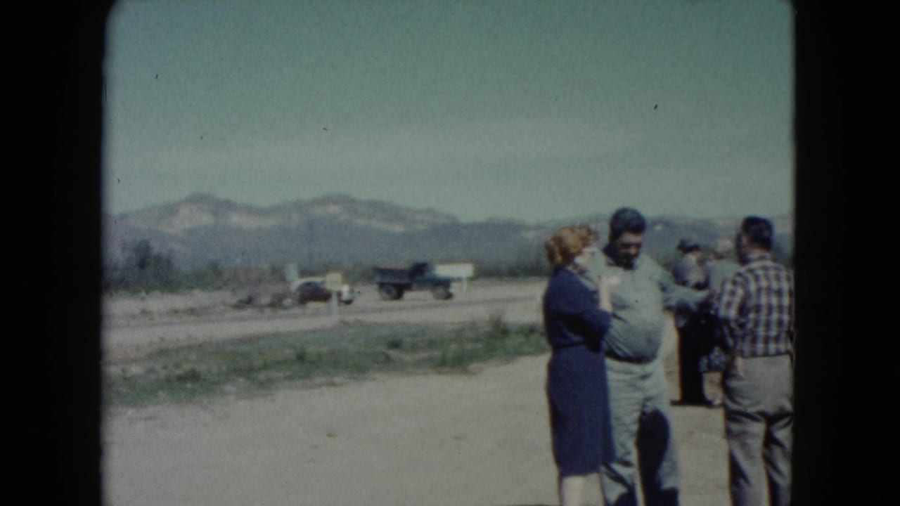 Adults stand talking on a dirt roadside with trucks and mountains behind them.