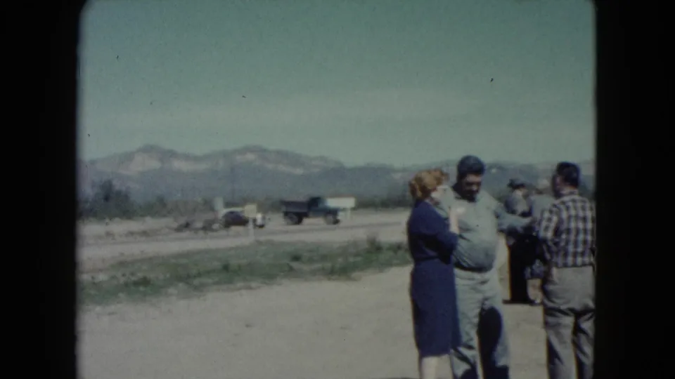 In Nevada home-movie footage from 1960, several adults appear to talk beside a dirt road with older vehicles and mountains in the background.