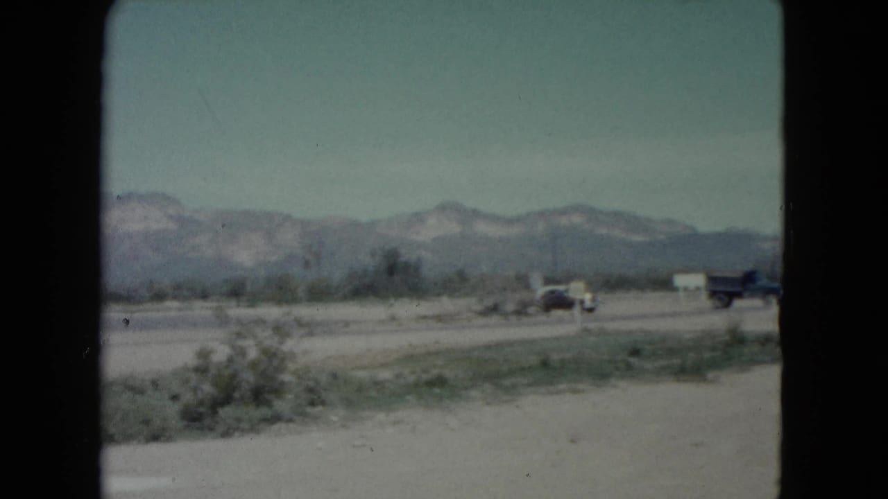 The camera looks across a Nevada roadside landscape with sparse scrub, distant mountains and older vehicles partly visible along the road.