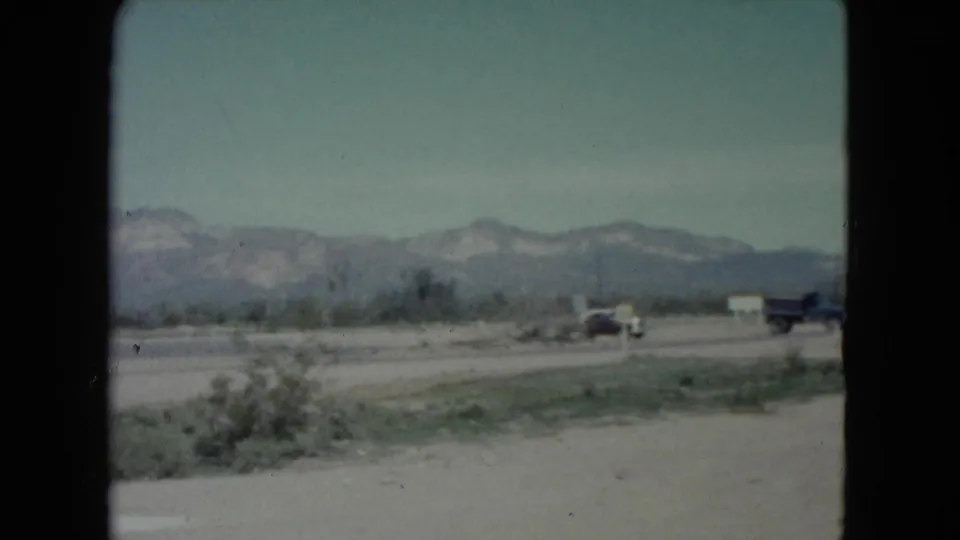 The camera looks across a Nevada roadside landscape with sparse scrub, distant mountains and older vehicles partly visible along the road.