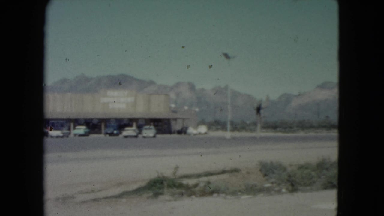 1960 Nevada home-movie frame showing a parking lot with cars before a long retail building and mountains in the background.