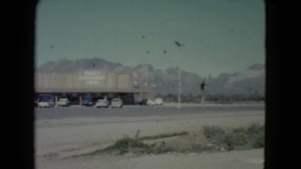 1960 Nevada home-movie frame showing a parking lot with cars before a long retail building and mountains in the background.